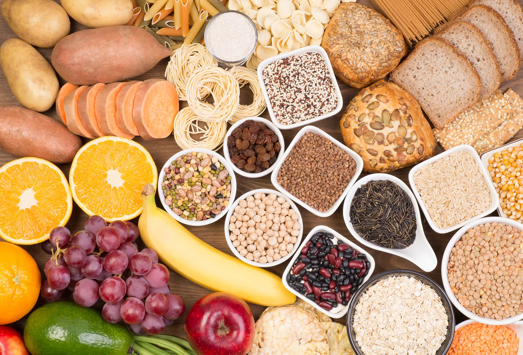 Diverse assortment of carbohydrate-rich staple foods on a wooden surface, including grains, pasta, bread, potatoes, sweet potatoes, legumes, and various fruits.