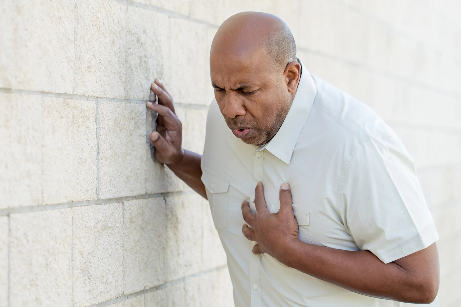 Adult man with a bald head clutching his chest in pain, leaning against a light-colored brick wall for support.