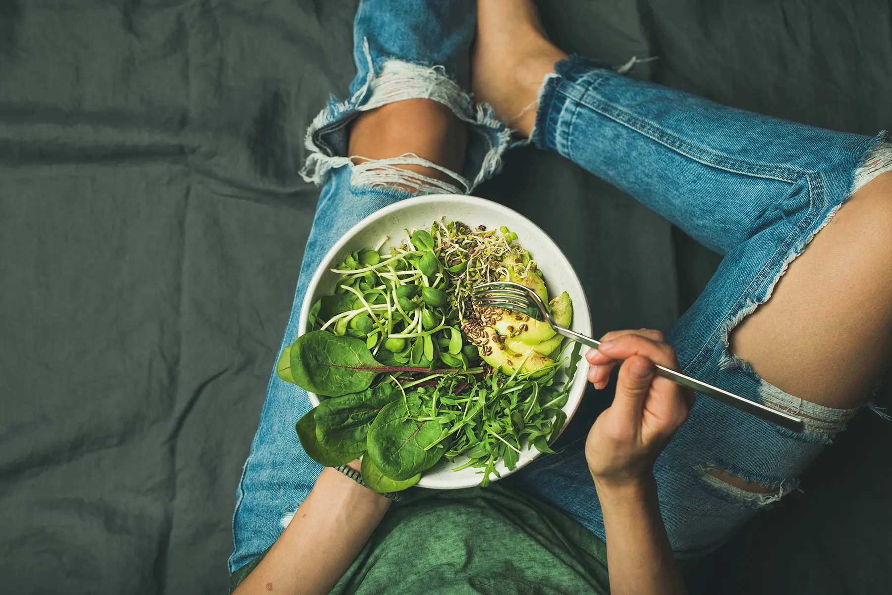 Overhead view of a person in ripped blue jeans holding a bowl of healthy salad with fresh greens, sprouts, sliced avocado, and seeds, on a dark gray surface.