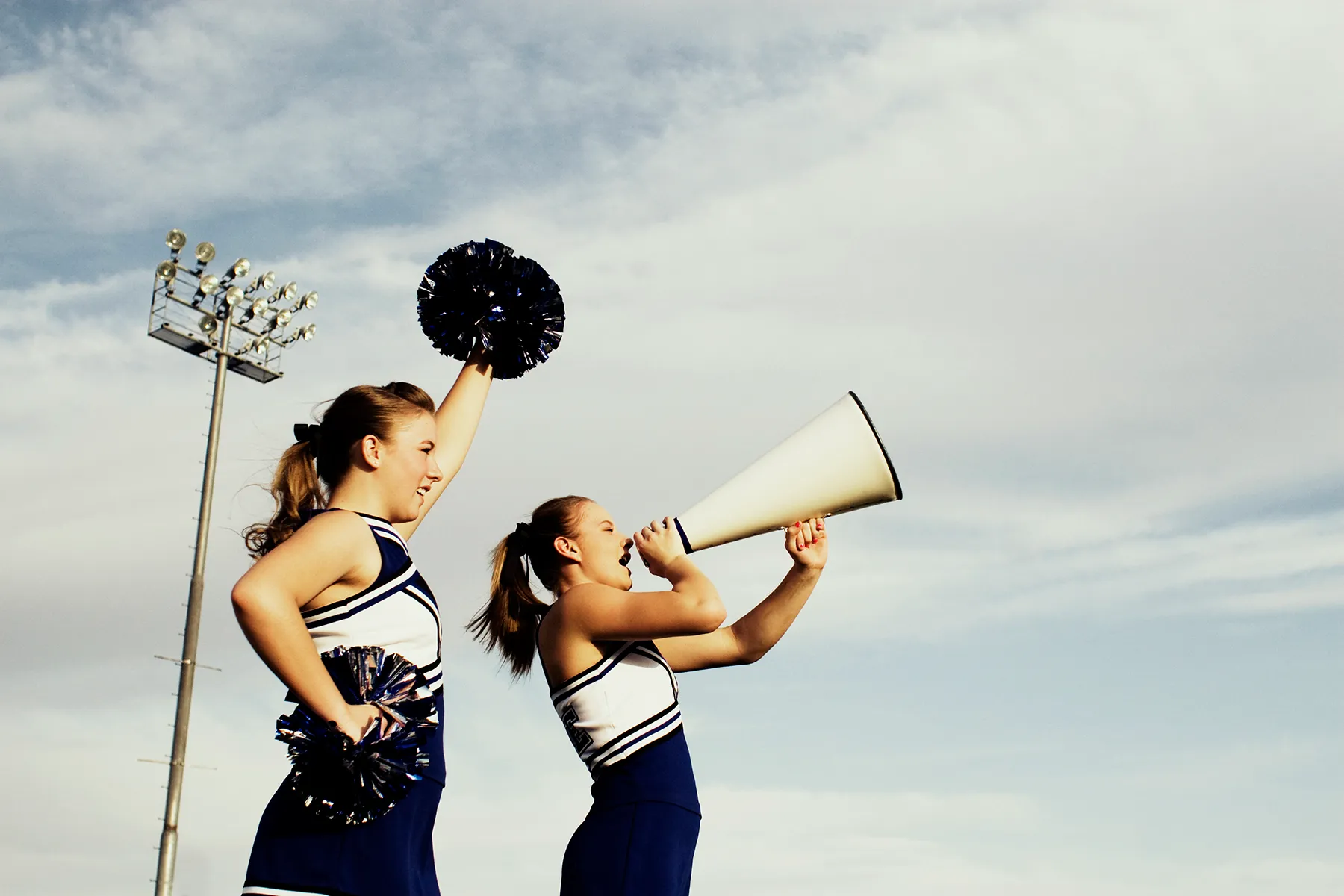 Two cheerleaders: one shouts enthusiastically into a white megaphone, while the other raises dark pom-poms. Both wear navy and white uniforms against a cloudy sky with a stadium light pole.