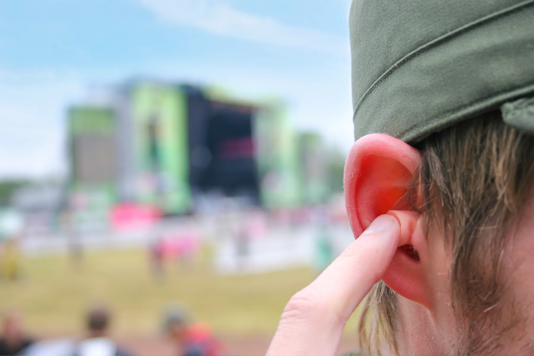 Person inserting an earplug into their right ear, wearing an olive-green cap, with a blurred outdoor music festival stage in the background.