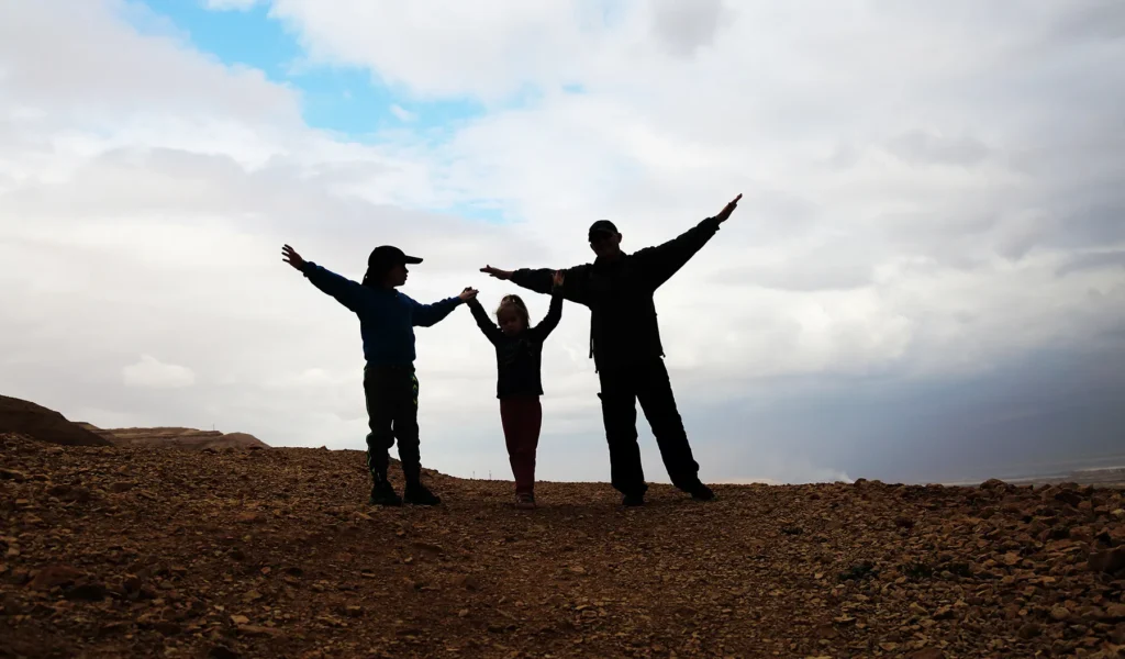 Family silhouette with outstretched arms on rocky terrain under a cloudy sky.