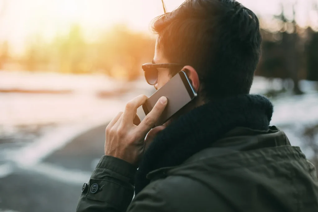 Rear view of a man talking on a smartphone, wearing sunglasses and a jacket, with warm golden hour sunlight in the background.