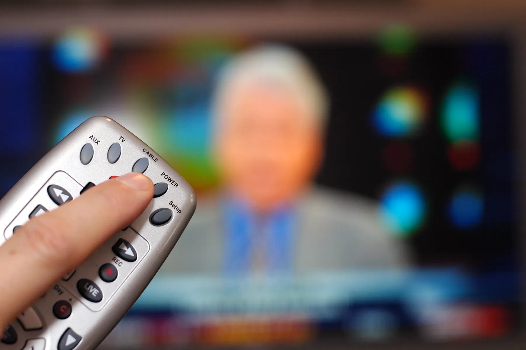 A hand presses the 'CABLE' button on a silver remote control, with a blurred television screen showing a person in the background.