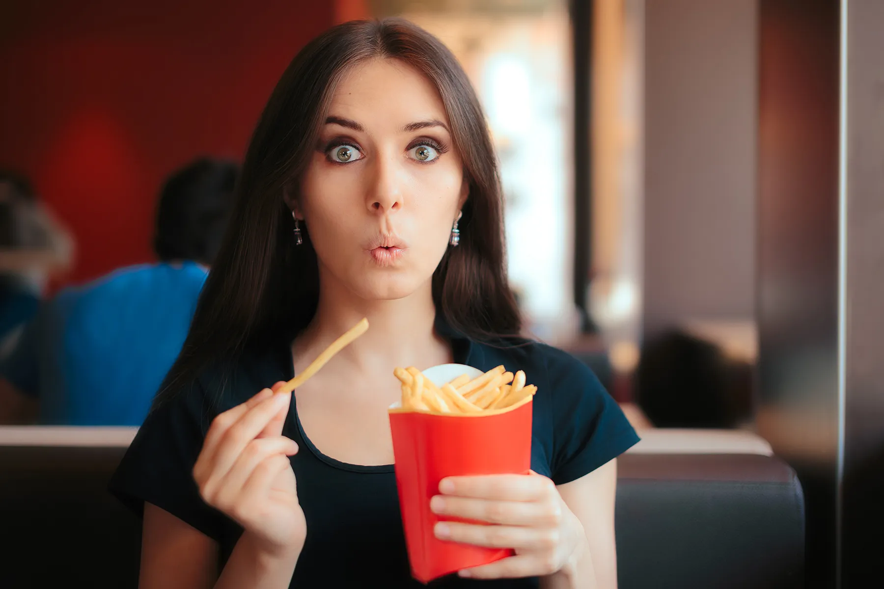 Young woman with wide, surprised eyes and pursed lips holding a single french fry and a red container of french fries in a casual dining or fast food setting.