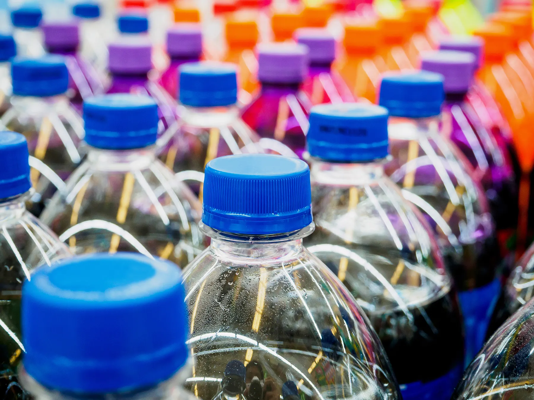 Rows of clear plastic beverage bottles with colorful caps, featuring prominent blue caps in the foreground and blurred purple, orange, and red caps in the background. Condensation and light reflections are visible on the cold bottles, suggesting a supermarket aisle.