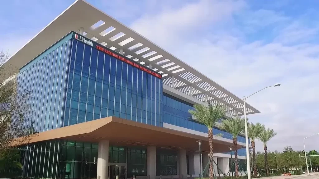 Modern UHealth Medical Centre building with a sleek blue glass facade and a distinctive white roof structure, under a sunny sky with palm trees in the foreground.