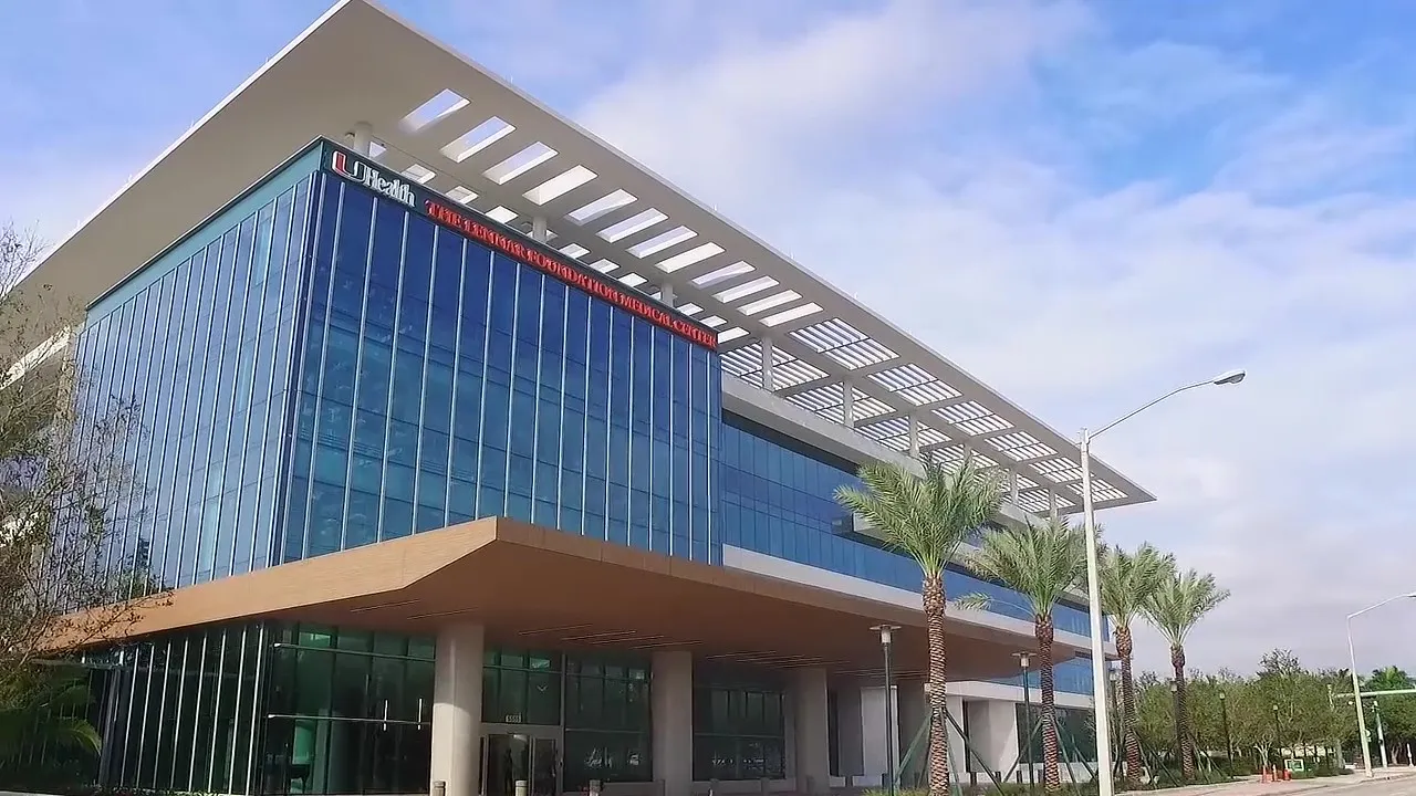 Modern UHealth Medical Centre building with a sleek blue glass facade and a distinctive white roof structure, under a sunny sky with palm trees in the foreground.