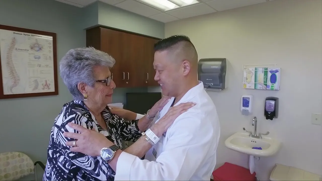 A male healthcare professional in a white coat and an elderly female patient with gray hair share a warm, supportive moment in a medical exam room, with a spine diagram and medical supplies visible in the background.