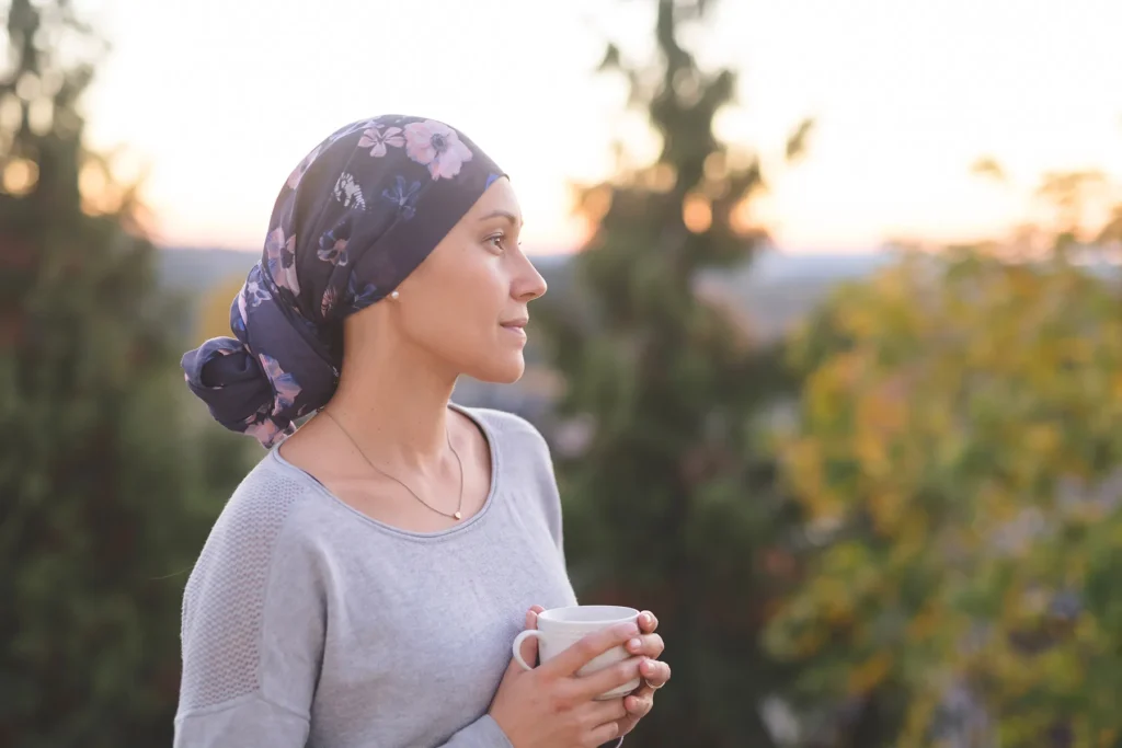 Woman in a dark blue floral turban-style headscarf and gray sweater, holding a white mug, looking serenely at a golden hour sunset over a blurred natural landscape.