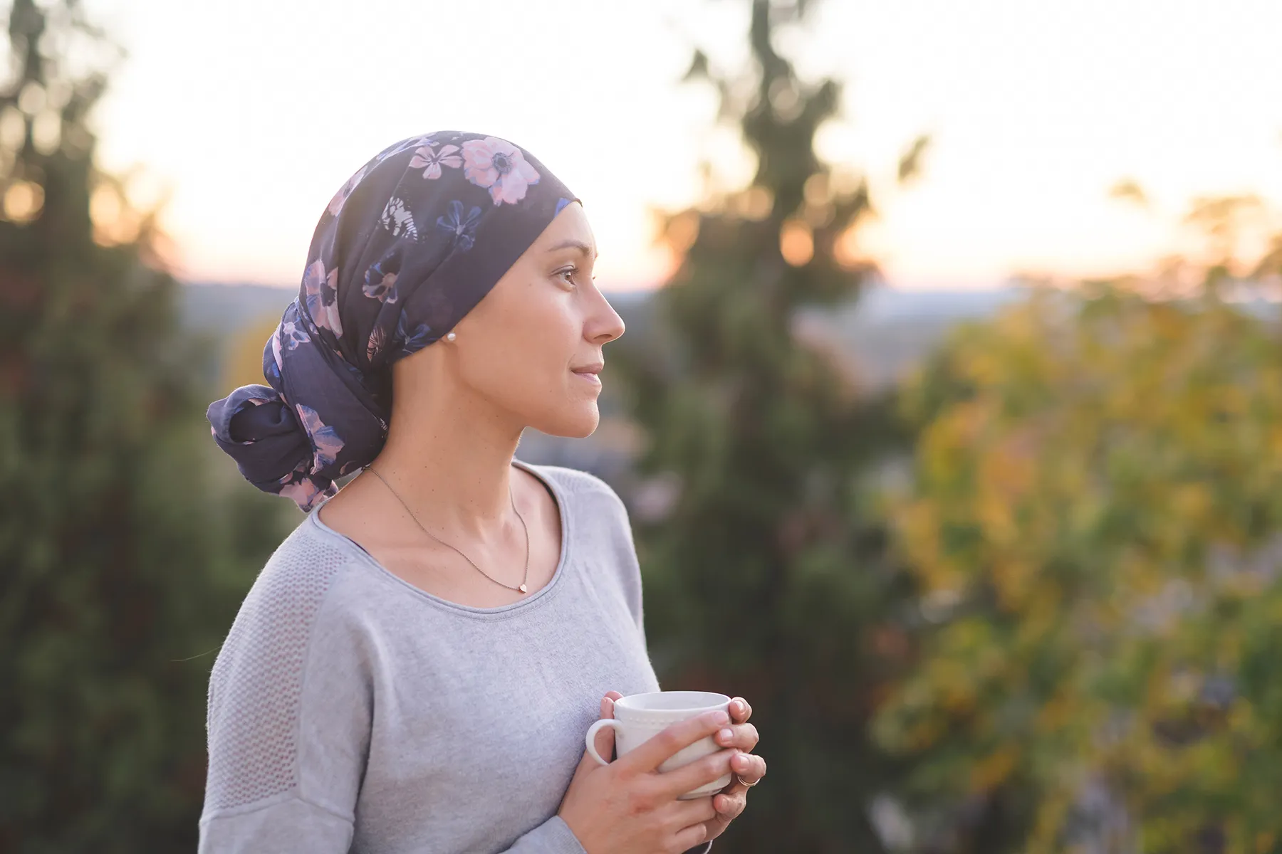 Woman in a dark blue floral turban-style headscarf and gray sweater, holding a white mug, looking serenely at a golden hour sunset over a blurred natural landscape.