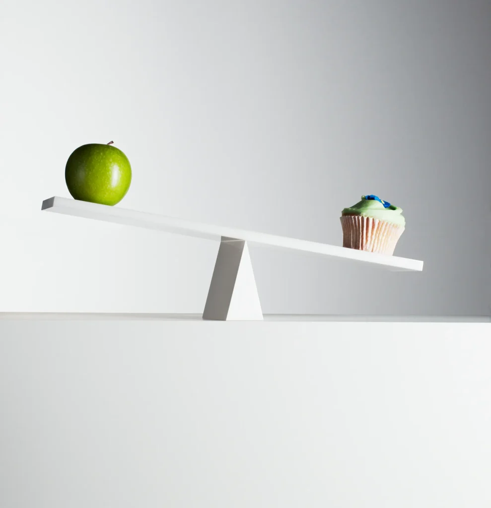 Green apple on a white balance scale, tilted lower than a cupcake with green frosting and blue sprinkles, illustrating a healthy choice versus a sweet treat.