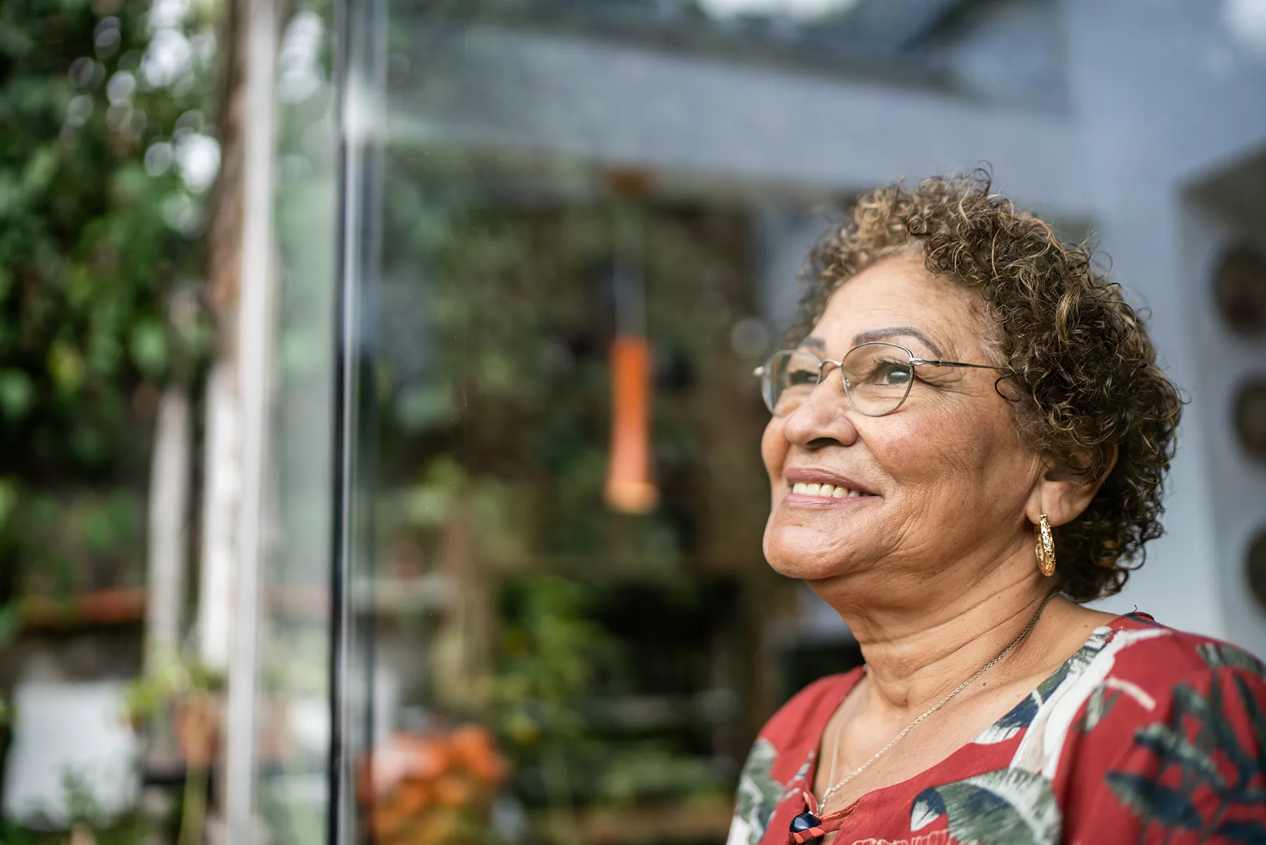 Smiling senior woman with curly hair and glasses, wearing a red patterned shirt, looking thoughtfully out a window with blurry green foliage reflections.