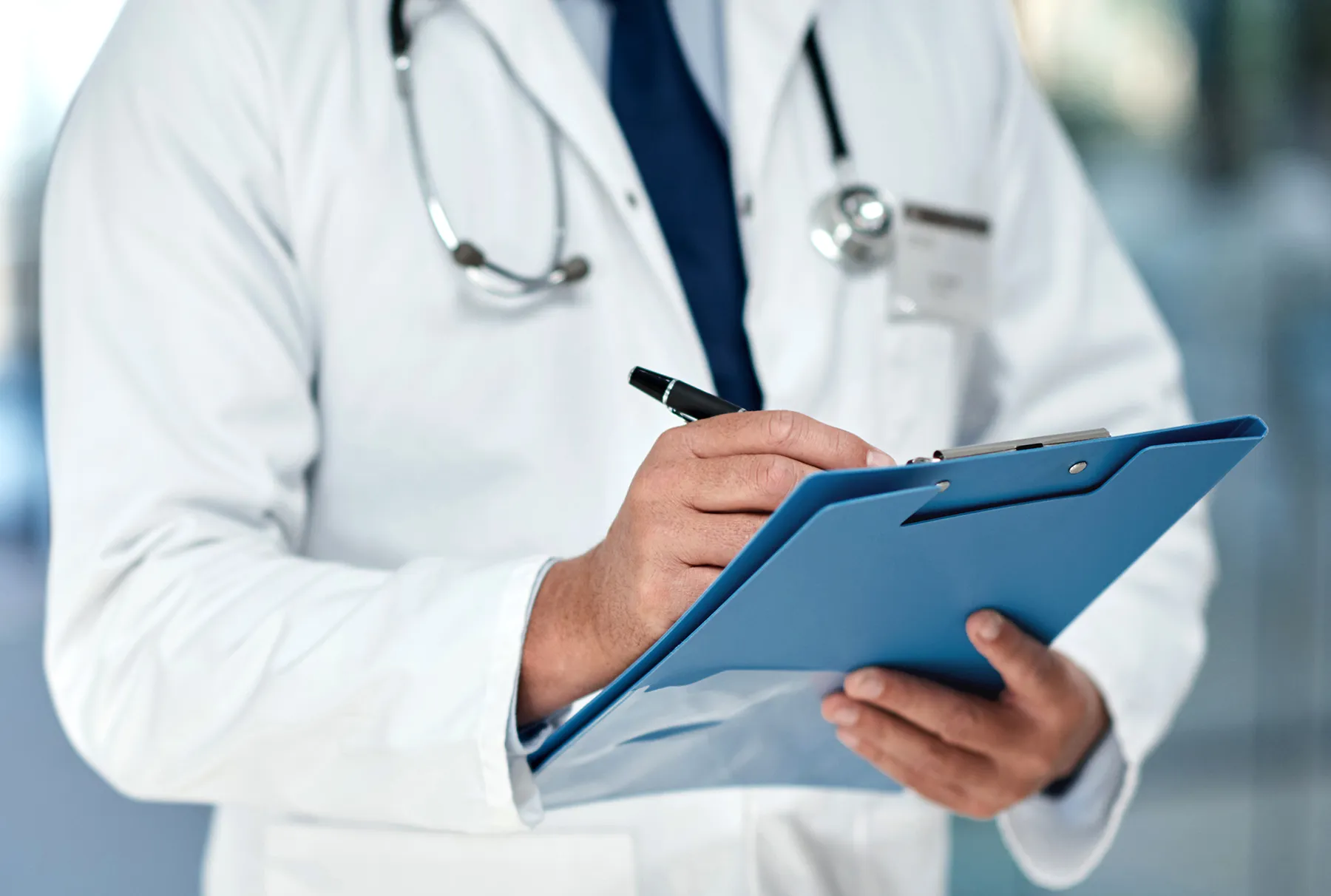 Medical professional in a white lab coat and stethoscope documenting on a blue clipboard with a pen.