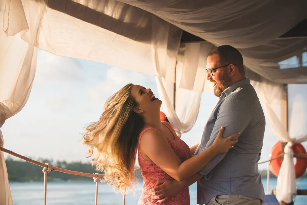 Joyful couple, the woman with long blonde hair and the man with a beard, embracing and laughing heartily on a sunny boat deck with draped fabric overhead, overlooking the water.