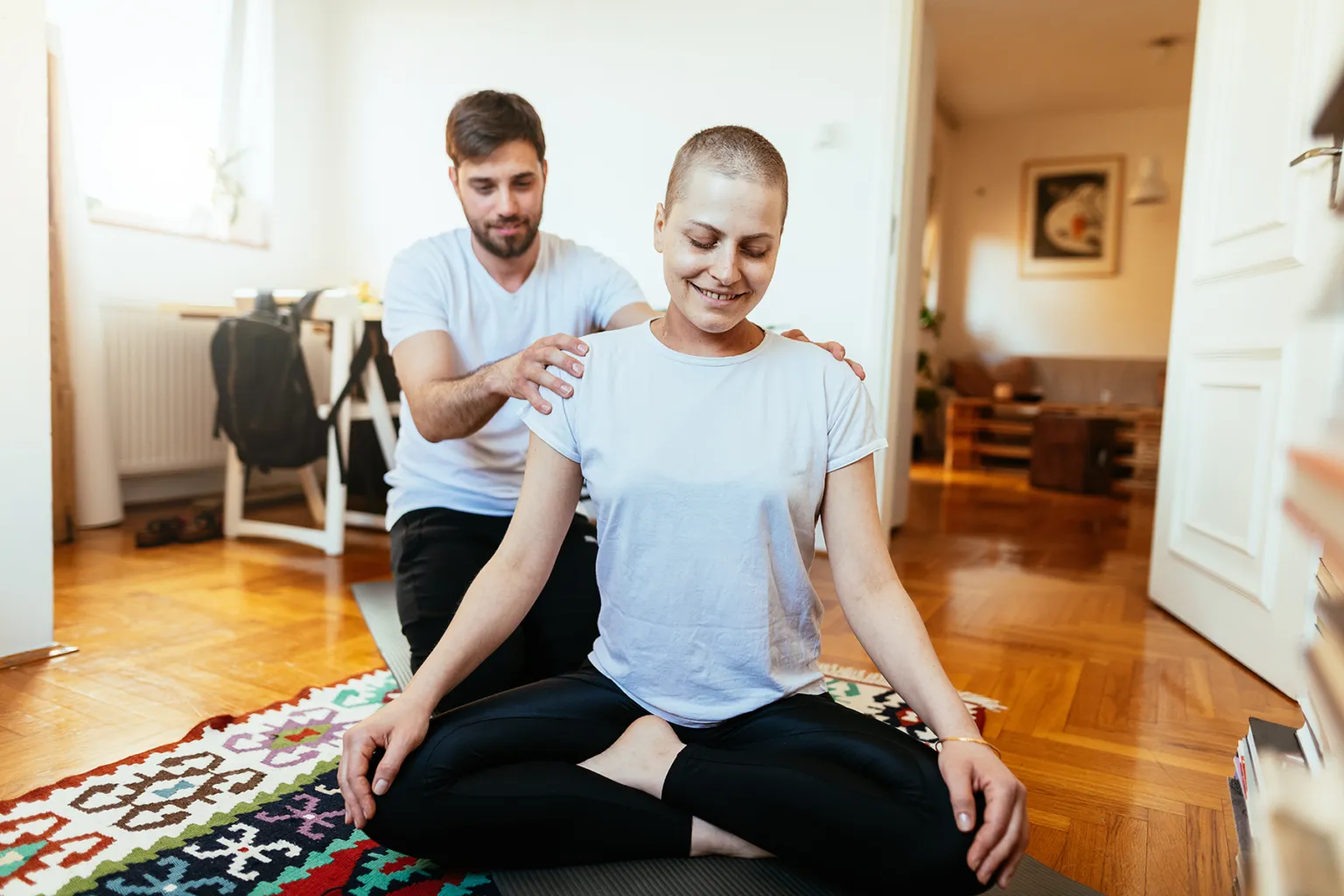 A woman with a shaved head meditating on a mat, receiving gentle shoulder support from a man in a bright home. Highlights wellness and care.