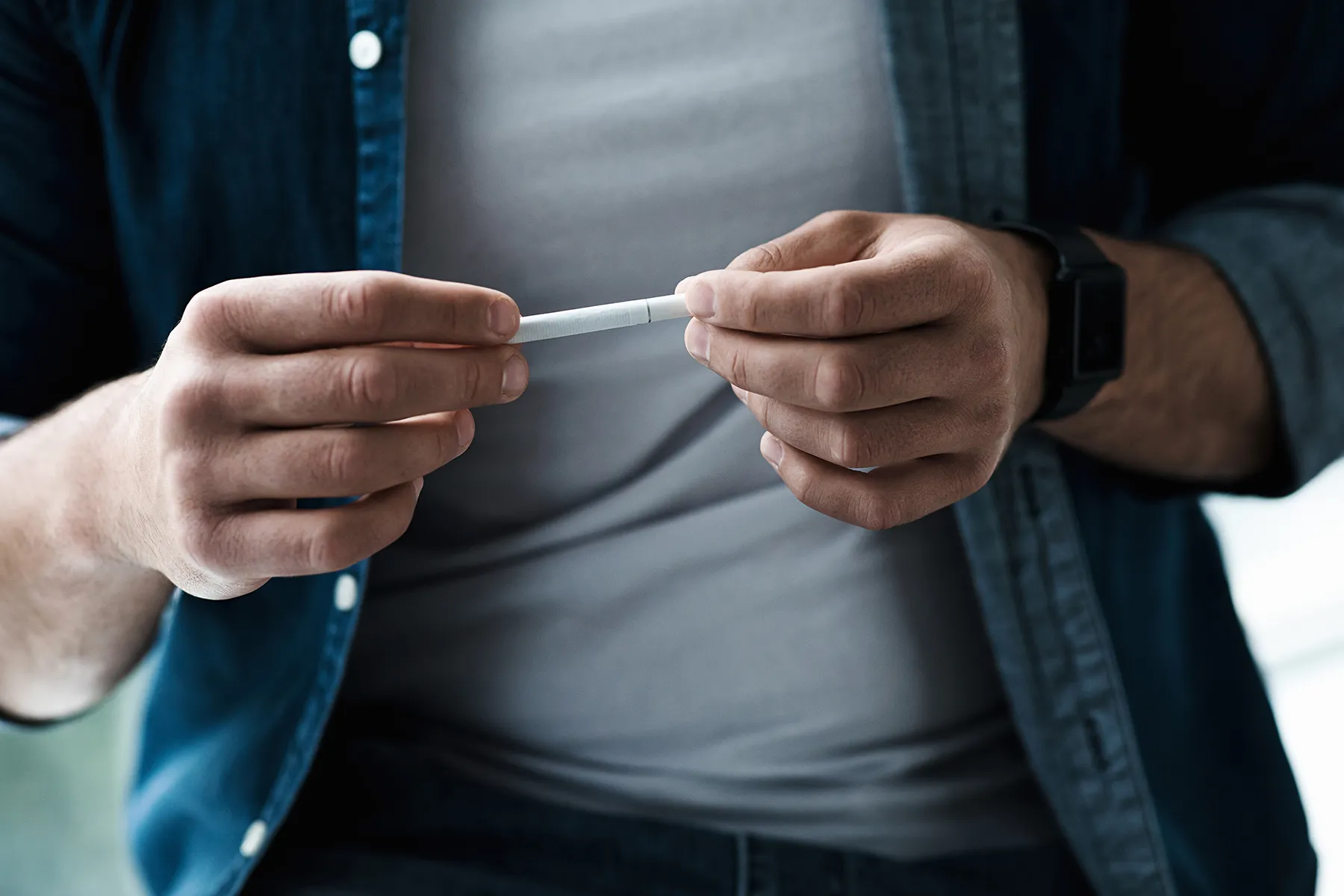 Person's hands holding and manipulating a white heat-not-burn tobacco stick. The person wears a blue denim shirt, a grey t-shirt, and a black fitness tracker on their wrist.