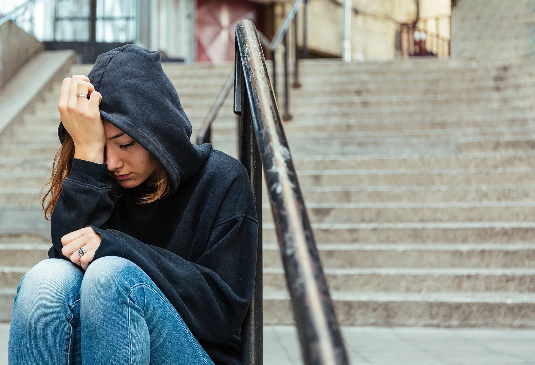 A young person in a black hoodie and jeans sits alone on concrete steps, head down and hand to forehead, conveying feelings of sadness, anxiety, or despair.