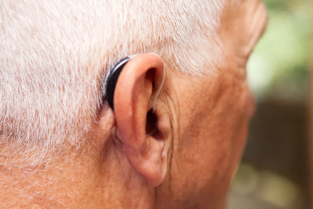 Close-up of an older adult's ear with a black behind-the-ear hearing aid, framed by white hair.
