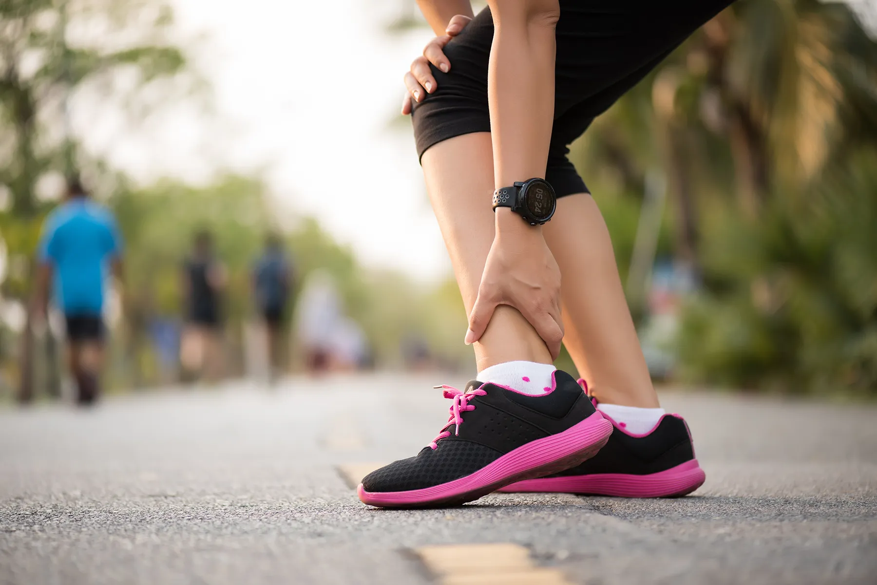 Runner clutching a painful knee and ankle on an outdoor running path, wearing black shorts and black and pink athletic shoes.