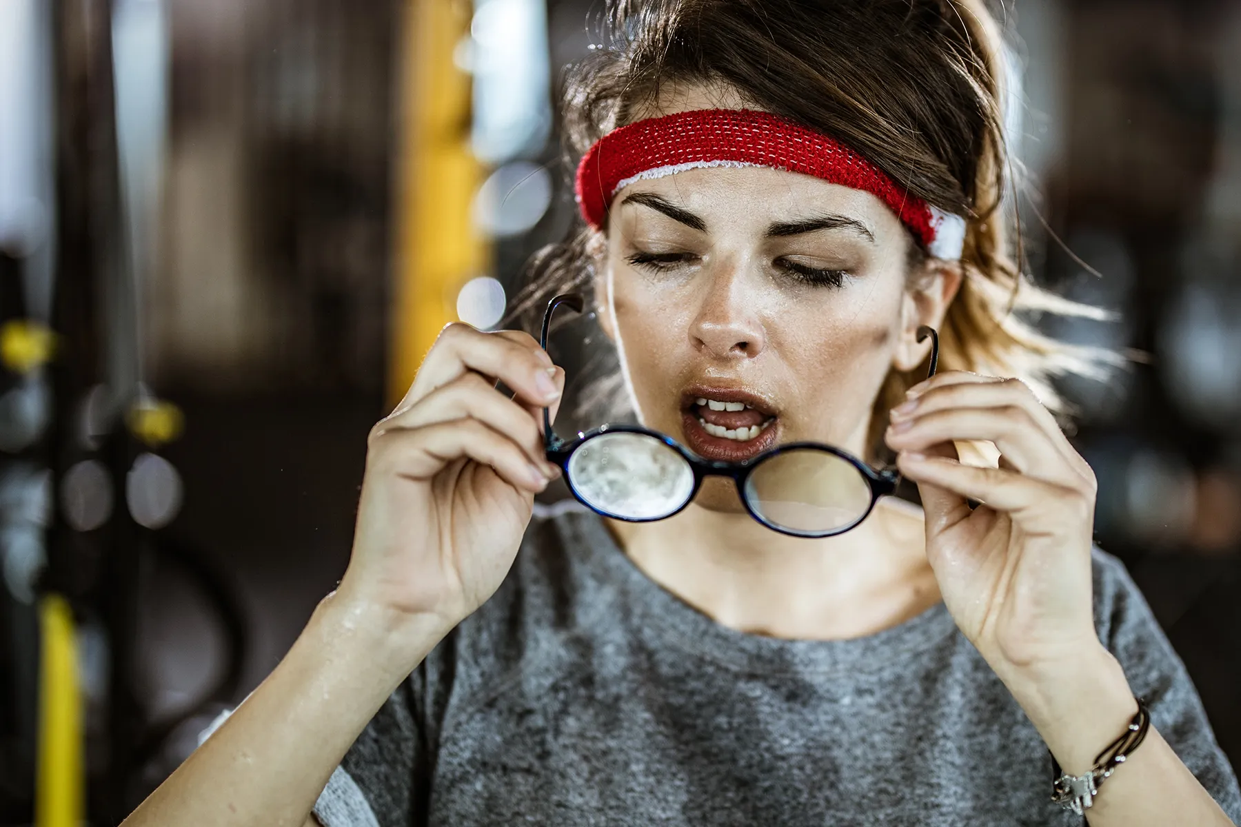 Exhausted and sweaty woman with a red headband holding fogged glasses after a gym workout.