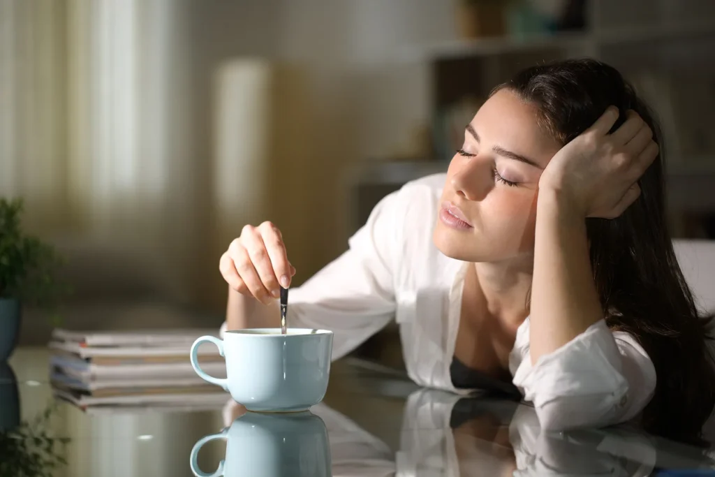 Sleepy young woman with closed eyes resting her head on her arm, stirring a light blue mug of coffee or tea on a glass table in the morning light.