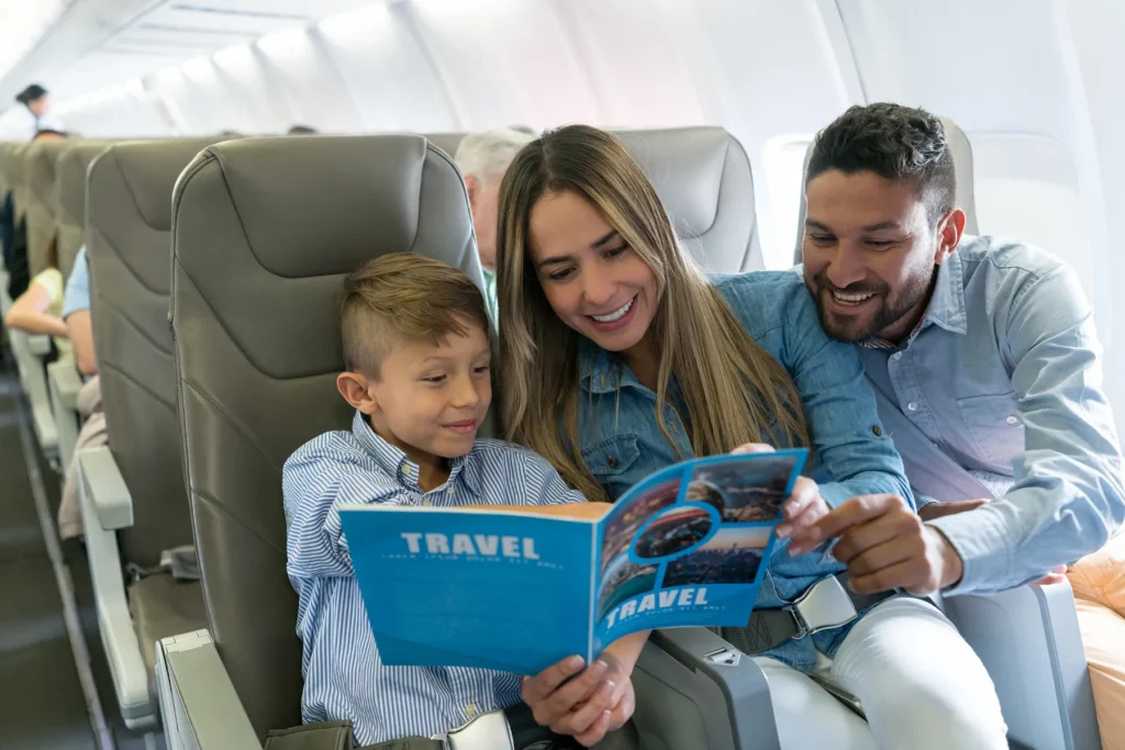 Smiling mother, father, and young son reading a blue "TRAVEL" magazine together on an airplane.