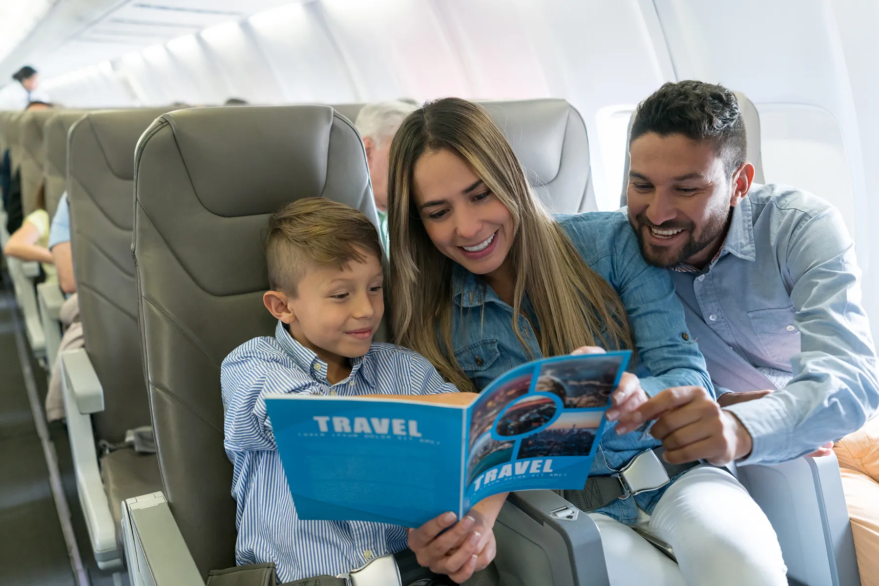 Smiling mother, father, and young son reading a blue "TRAVEL" magazine together on an airplane.
