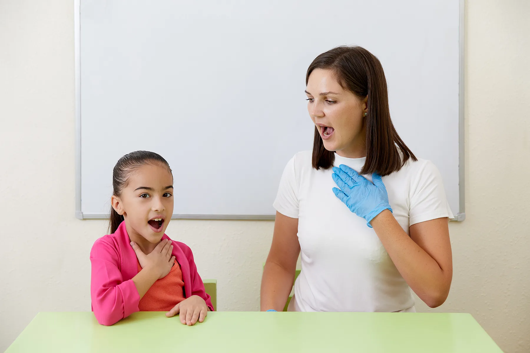 Speech therapist in a blue glove demonstrates vocal exercises to a young girl, both with hands on their chests and mouths open, in front of a whiteboard.