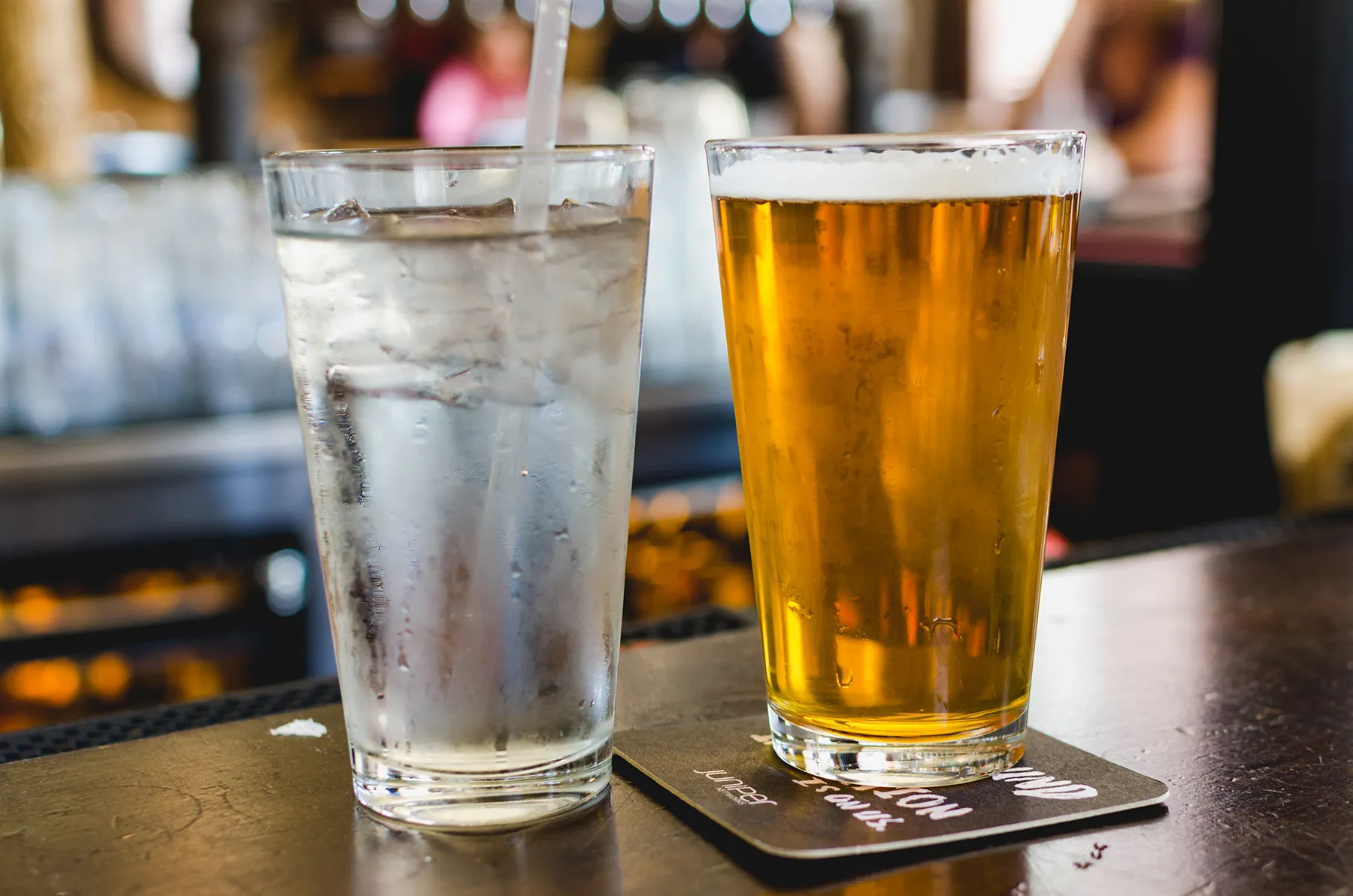 A cold glass of water with ice and a pint of golden beer sitting on a dark bar counter, with a blurred background of a bar interior.