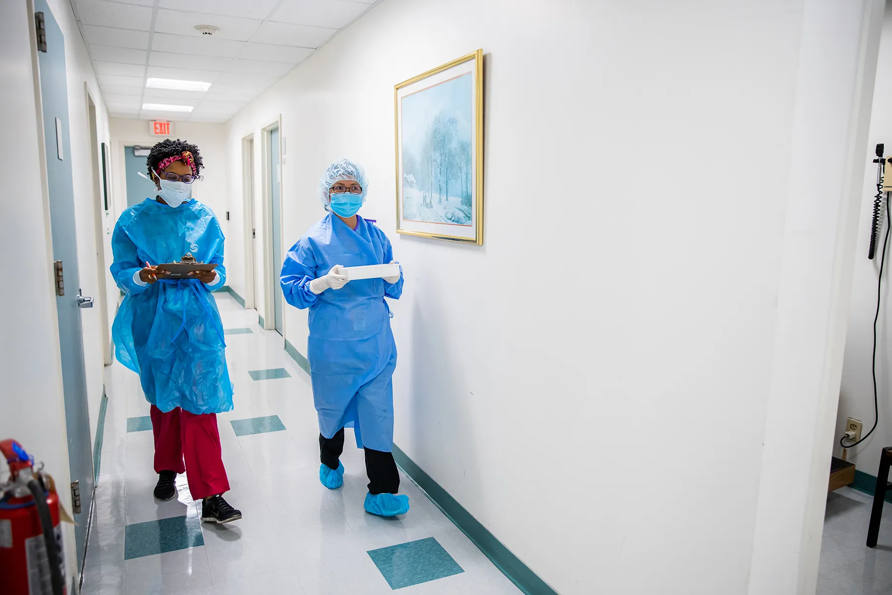 Two healthcare workers in blue protective gowns and masks walk down a well-lit hospital hallway. The worker on the left holds a clipboard, and the one on the right carries a white tray.