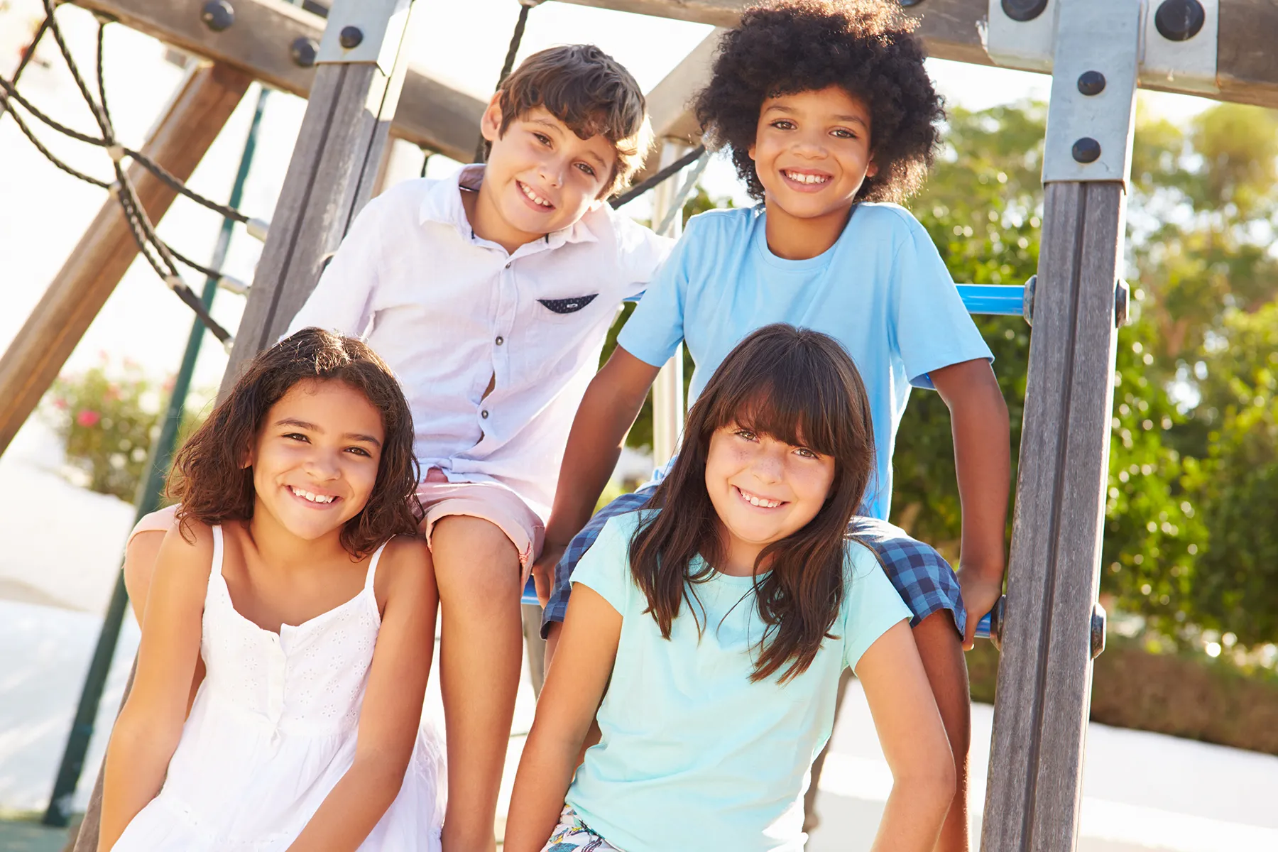 Four happy, diverse children smiling on a playground climbing structure outdoors.