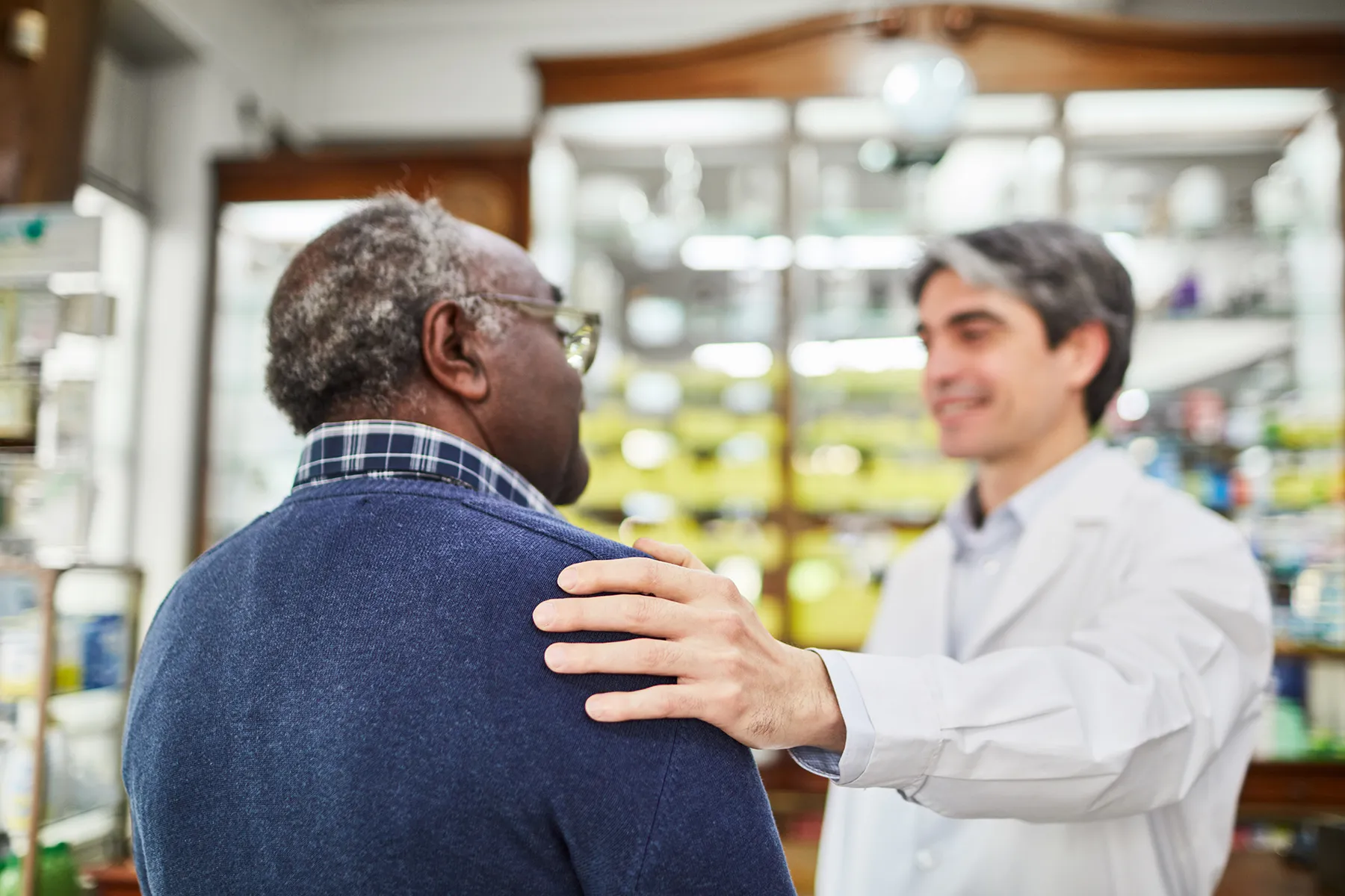 Male pharmacist places a reassuring hand on an older Black patient's shoulder in a pharmacy.