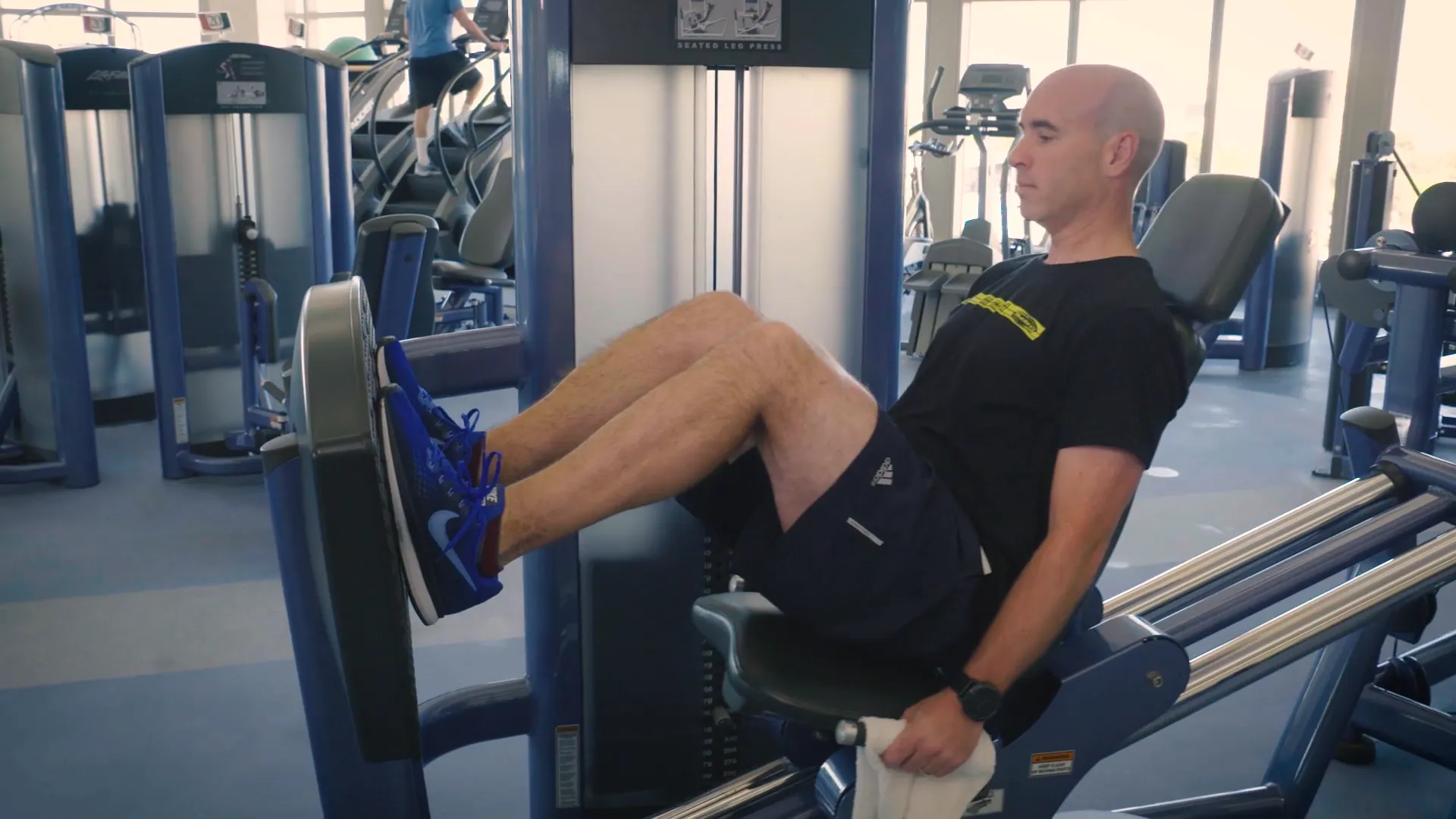 Man performing leg presses on a seated leg press machine in a bright gym. He wears a black t-shirt, dark shorts, and blue athletic shoes, focused on his workout.