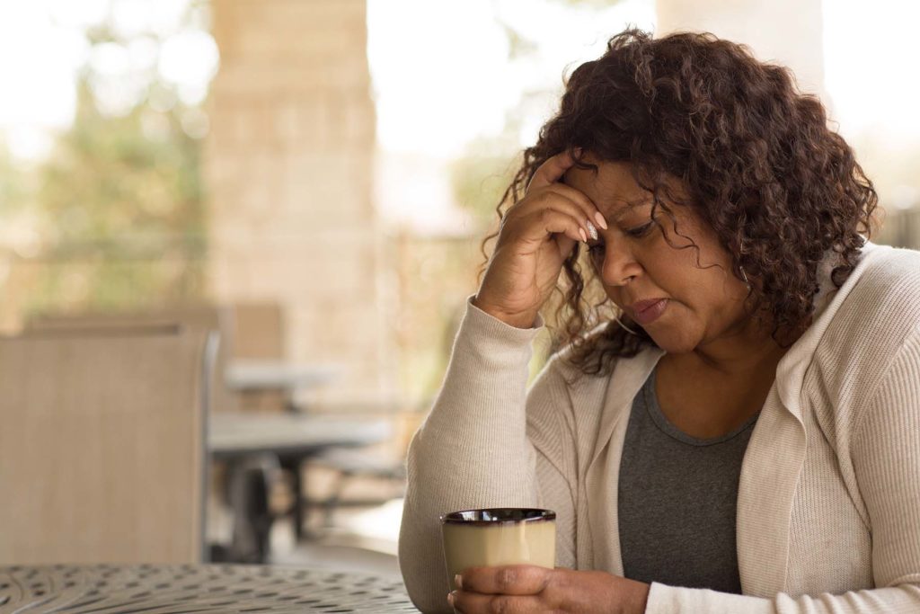 Older Black woman sits at table outside, head in hand