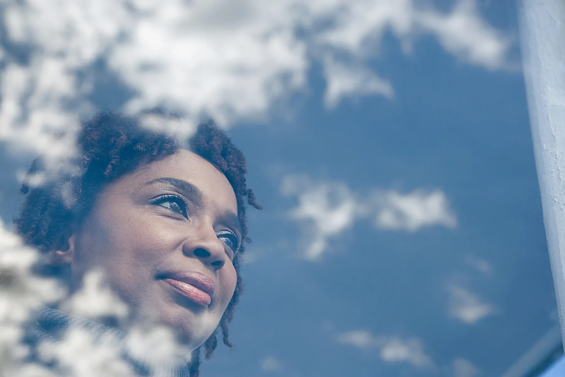 Woman with dark, curly hair looking upwards with a contemplative smile, reflections of a bright blue sky and white clouds overlaid on the scene.