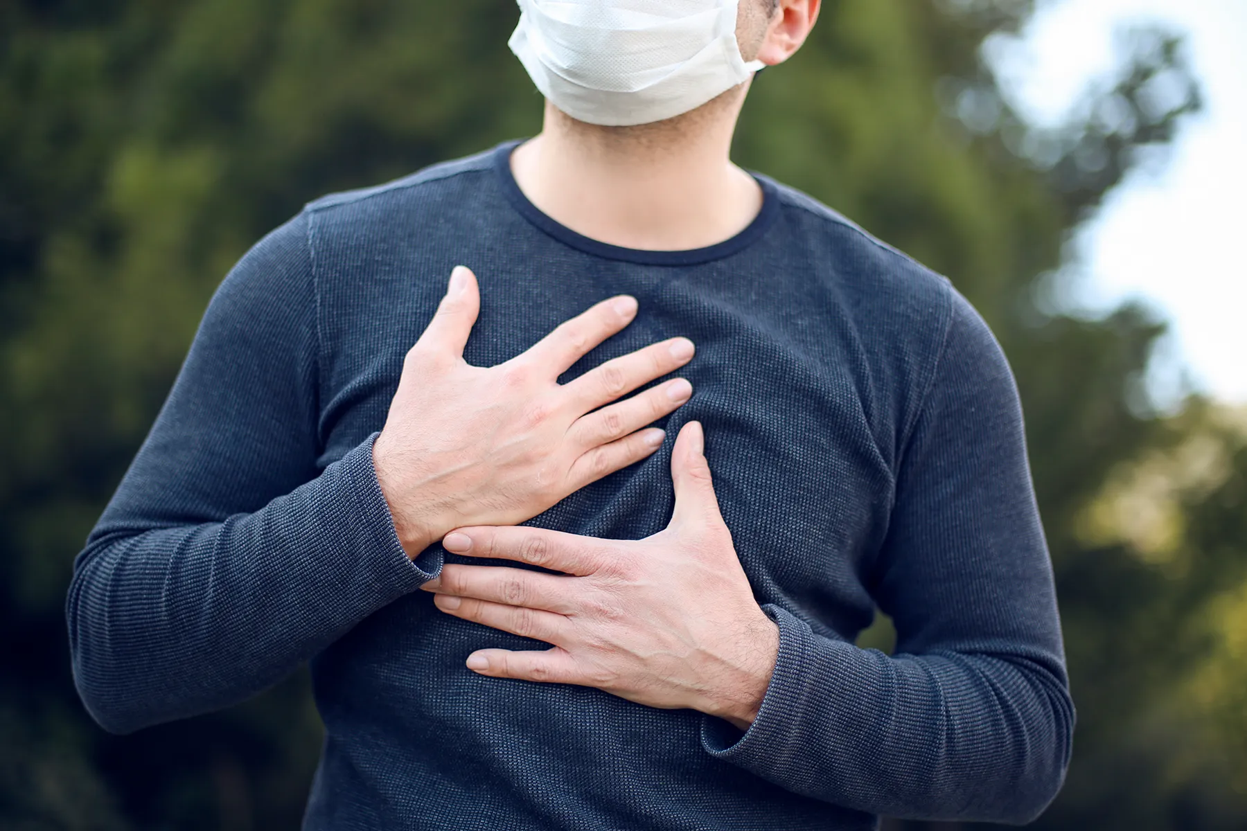 Man in a white face mask clutching his chest, suggesting chest pain or breathing difficulty, outdoors.