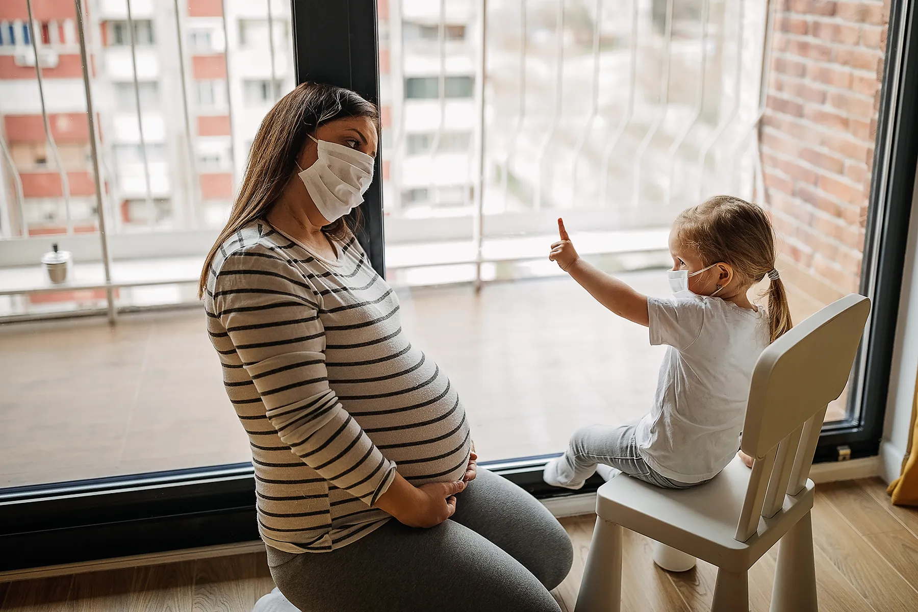 Pregnant woman and young child, both wearing face masks, sitting by a large window. The child points outside as the expectant mother holds her belly, reflecting a scene of family life during quarantine or a pandemic.