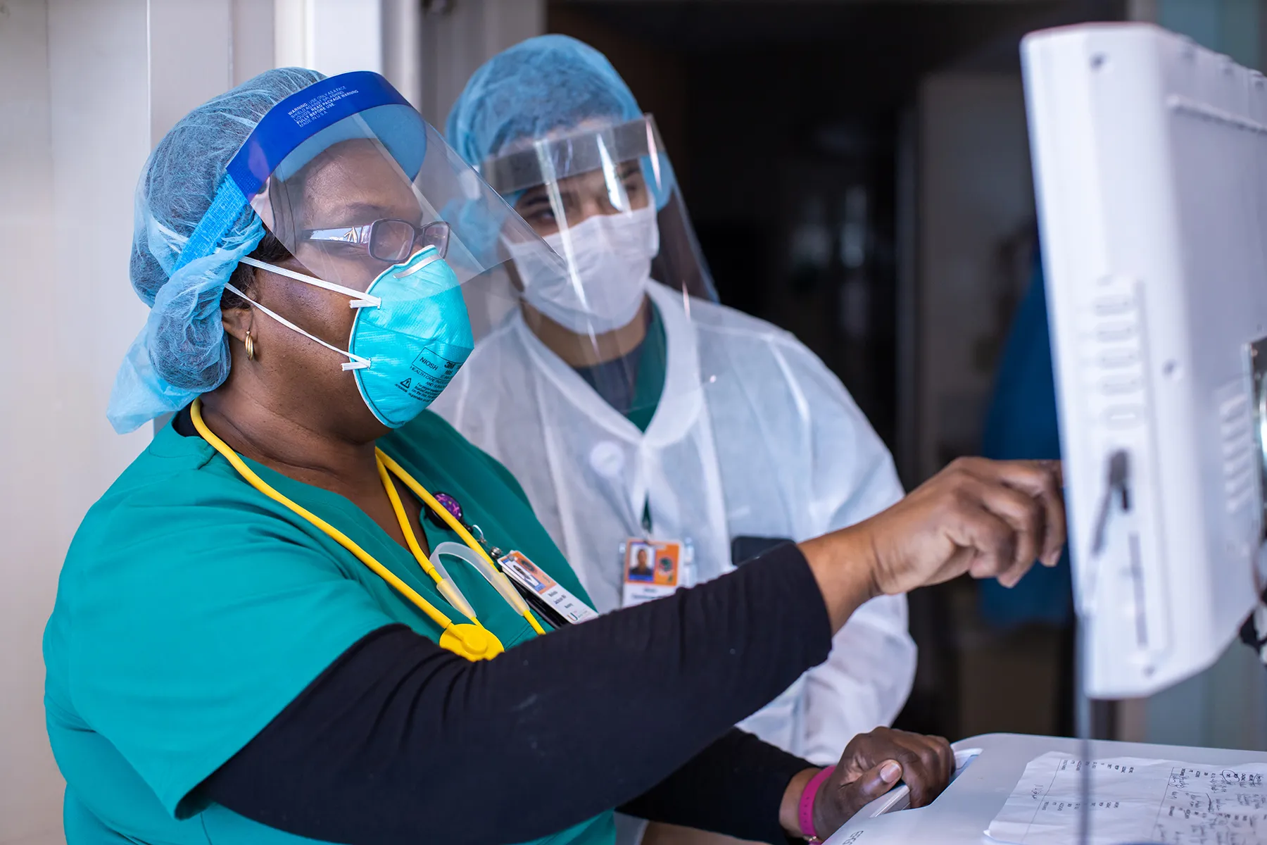 Two healthcare professionals in full personal protective equipment, including N95 masks and face shields, collaborating while looking at a computer screen in a medical facility. One person points at the monitor.