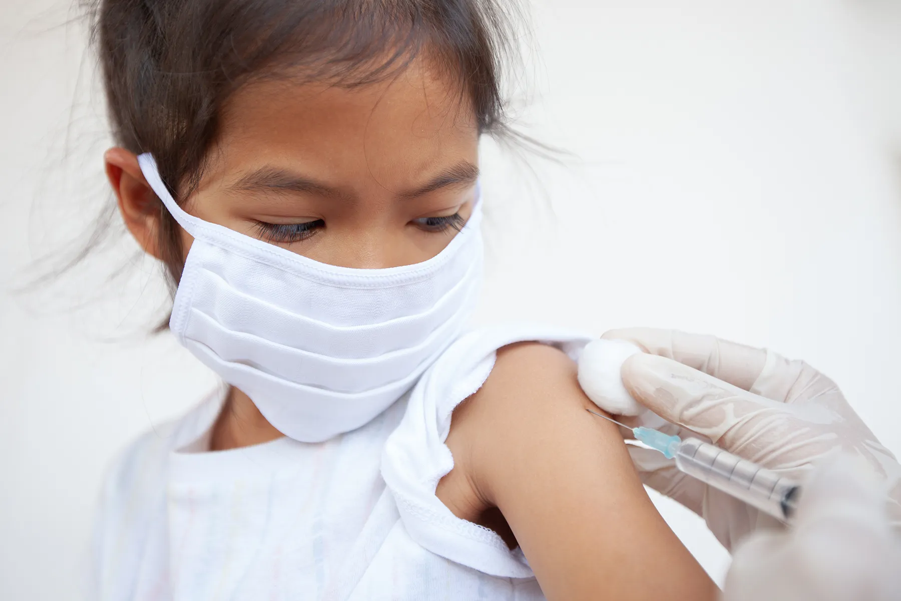 Young child in a white face mask receiving a vaccine injection in the arm.