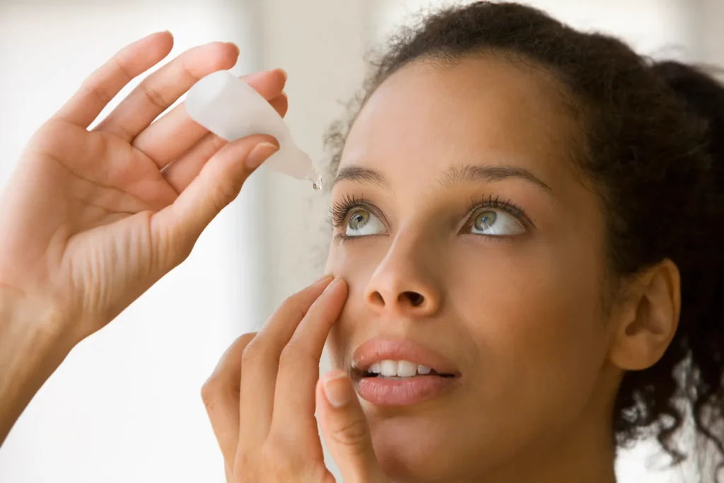 Woman applying eye drops, holding the bottle above her eye as a drop falls, while gently pulling down her lower eyelid.