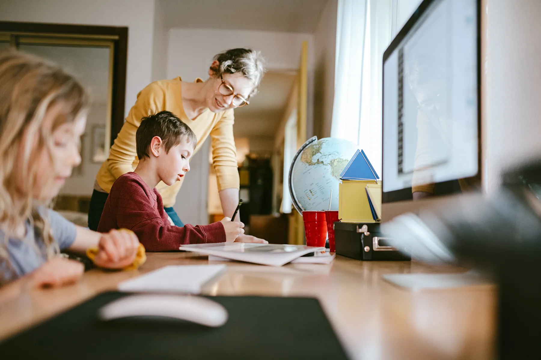 Mother assisting two children, a boy and a girl, with homeschooling at a wooden table. The boy writes in a notebook while the woman supervises. A globe, colorful geometric shapes, and a computer monitor are visible in the home learning environment.