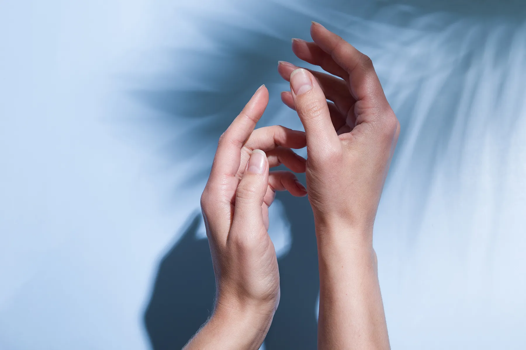 A pair of hands with smooth, healthy skin and natural nails, gently posed against a serene light blue background with soft palm leaf shadows.