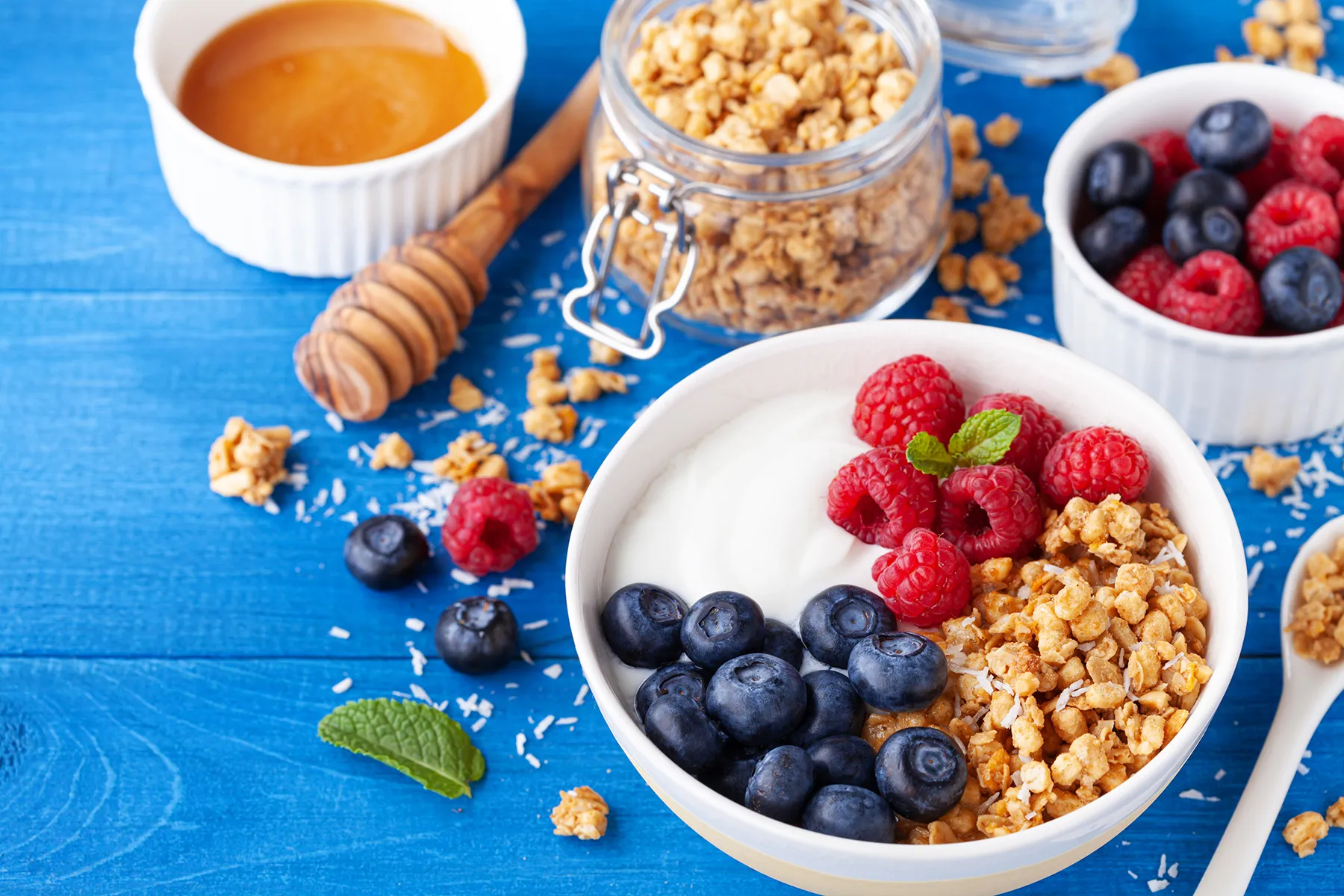 Healthy breakfast bowl with creamy yogurt, crunchy granola, fresh raspberries, and blueberries on a rustic blue wooden table. A jar of granola, honey, and additional berries are in the background.