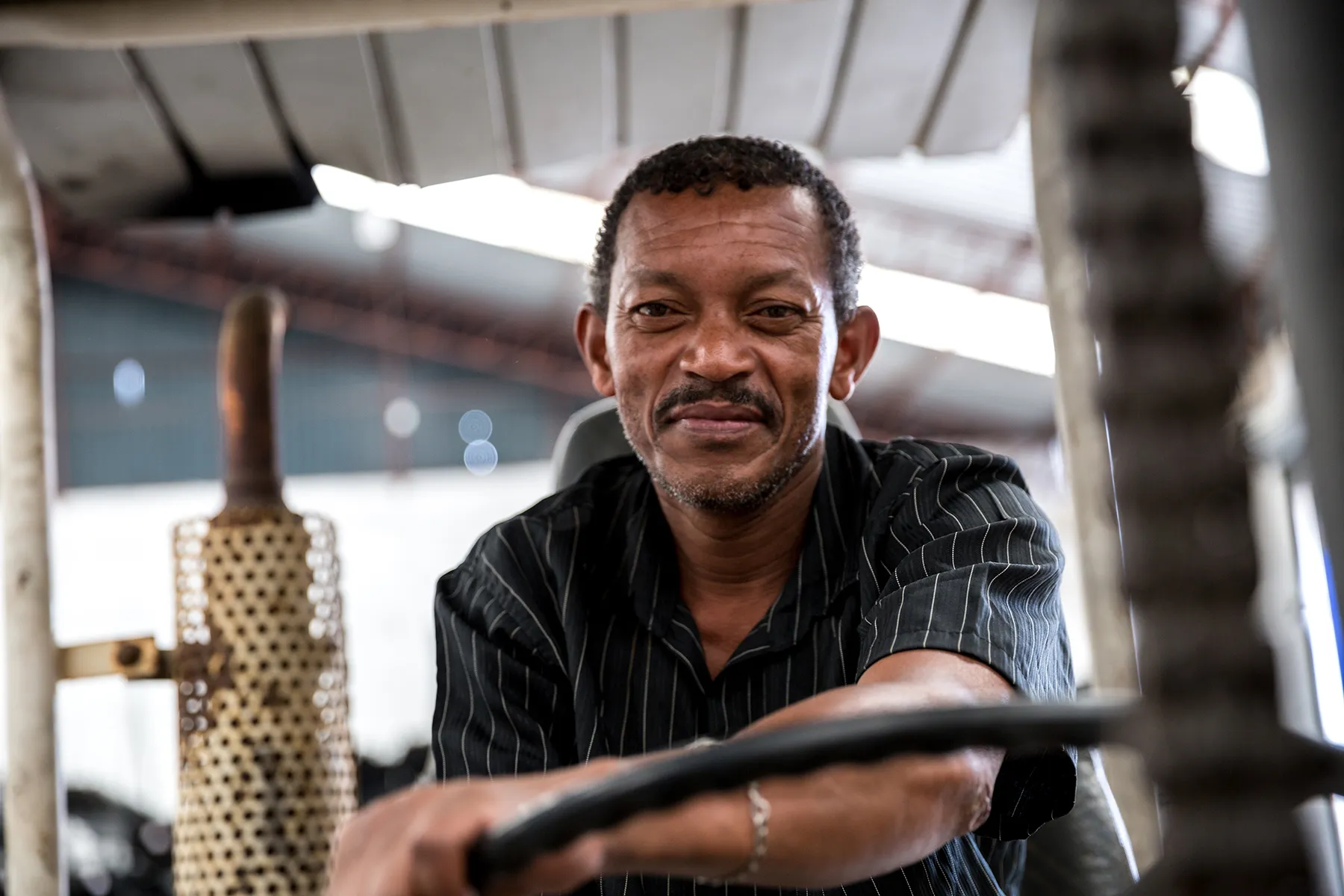 Smiling African American man with a mustache and goatee, wearing a pinstriped shirt, seated behind the wheel of heavy equipment. A blurred industrial interior with an exhaust pipe and a metal ceiling is in the background.