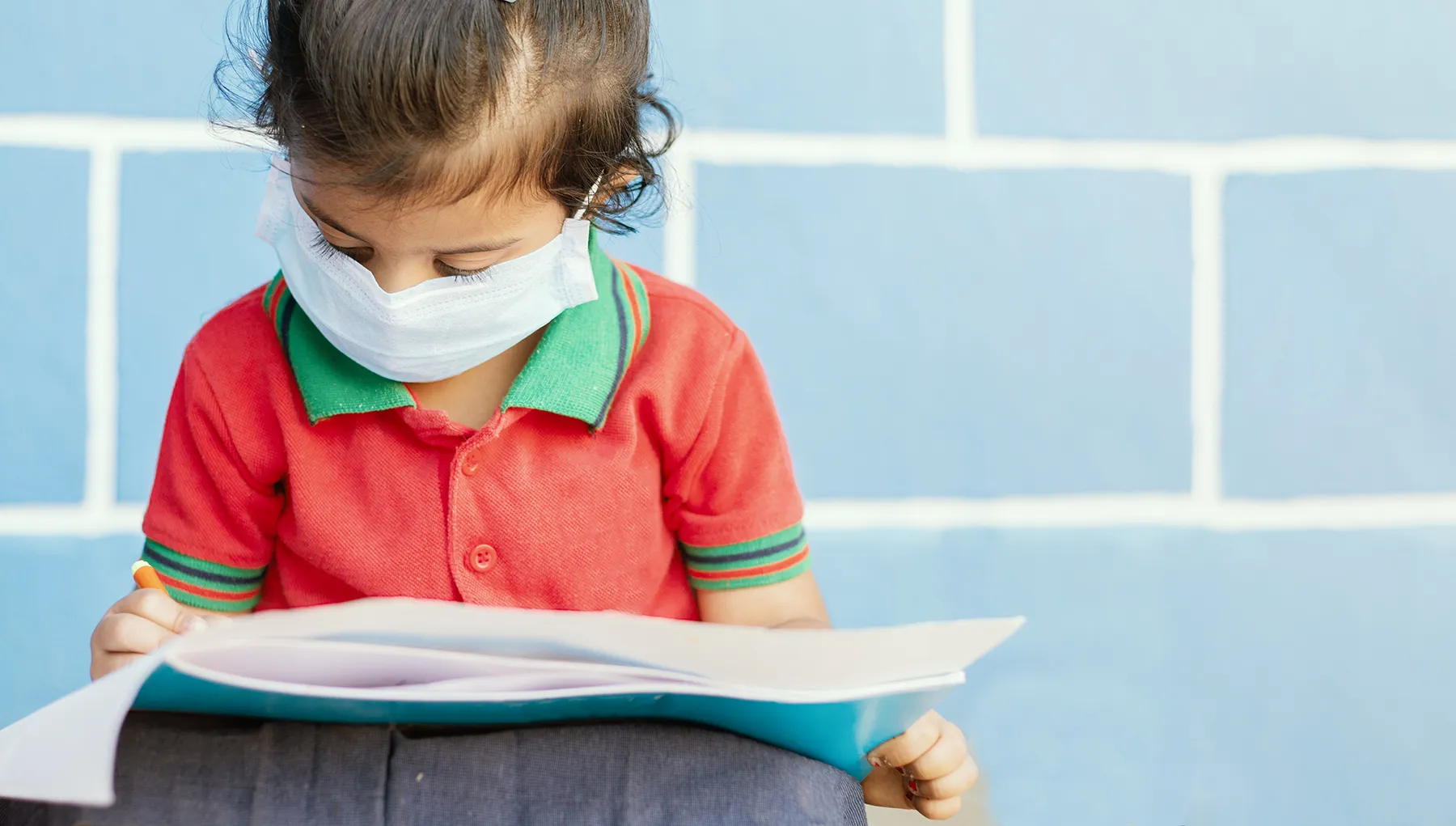 Young child wearing a white face mask, dressed in a red polo shirt, focused on writing or drawing on papers with a colorful marker, against a blurred light blue tiled wall.
