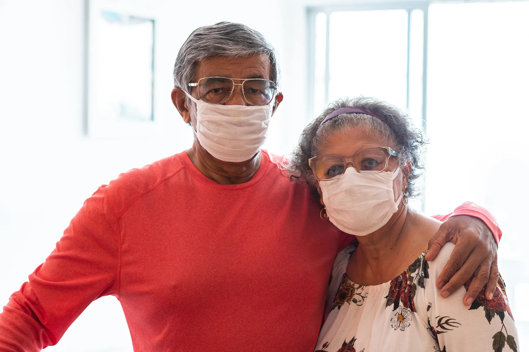 Elderly couple with grey hair wearing white face masks and glasses, embracing.