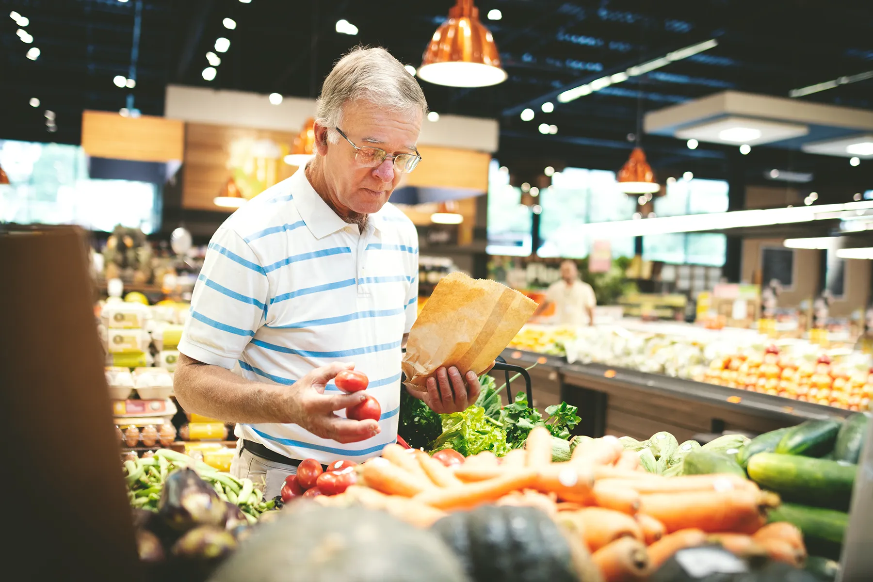 Elderly man in a supermarket's produce aisle, selecting fresh tomatoes and holding a brown paper bag.
