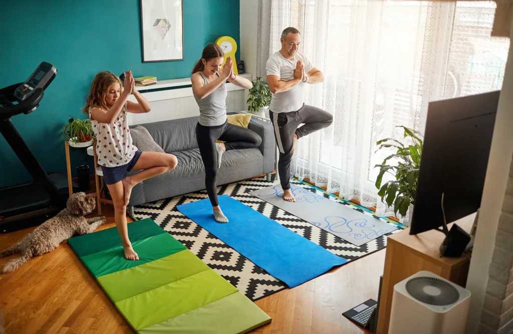 Father and daughters practicing yoga tree pose at home, with their dog lying on the floor.