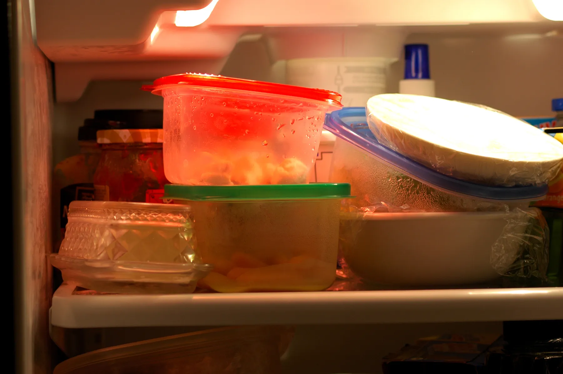 An interior view of a well-lit refrigerator shelf filled with various plastic food storage containers and bowls of leftovers, some showing condensation.